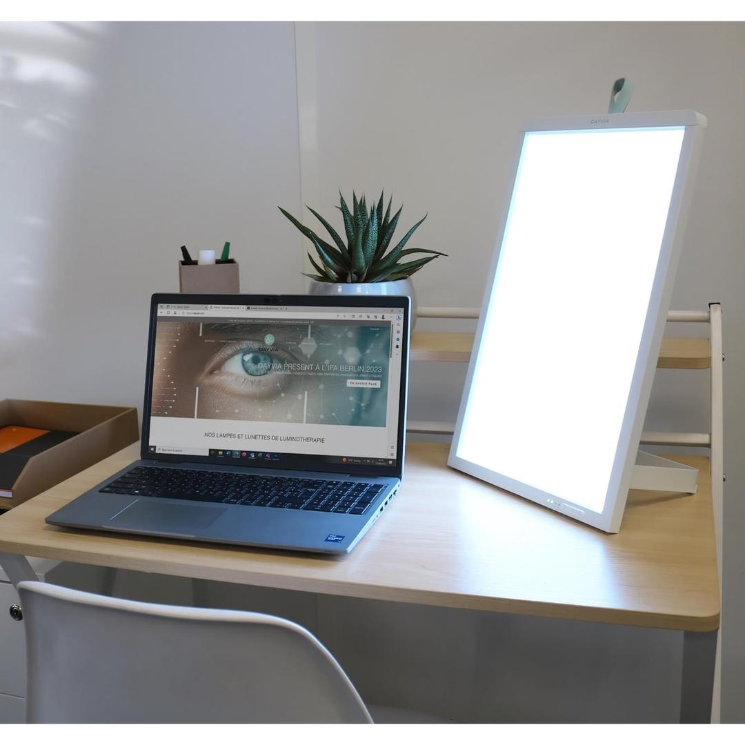 Lampe de luminothérapie LED, à poser ou à accrocher au mur Dayvia WELLBI UNILUX - Lampe de luminothérapie Dayvia WELLBI allumée sur un bureau, à côté d'un ordinateur portable