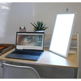 Lampe de luminothérapie LED, à poser ou à accrocher au mur Dayvia WELLBI UNILUX - Lampe de luminothérapie Dayvia WELLBI allumée sur un bureau, à côté d'un ordinateur portable