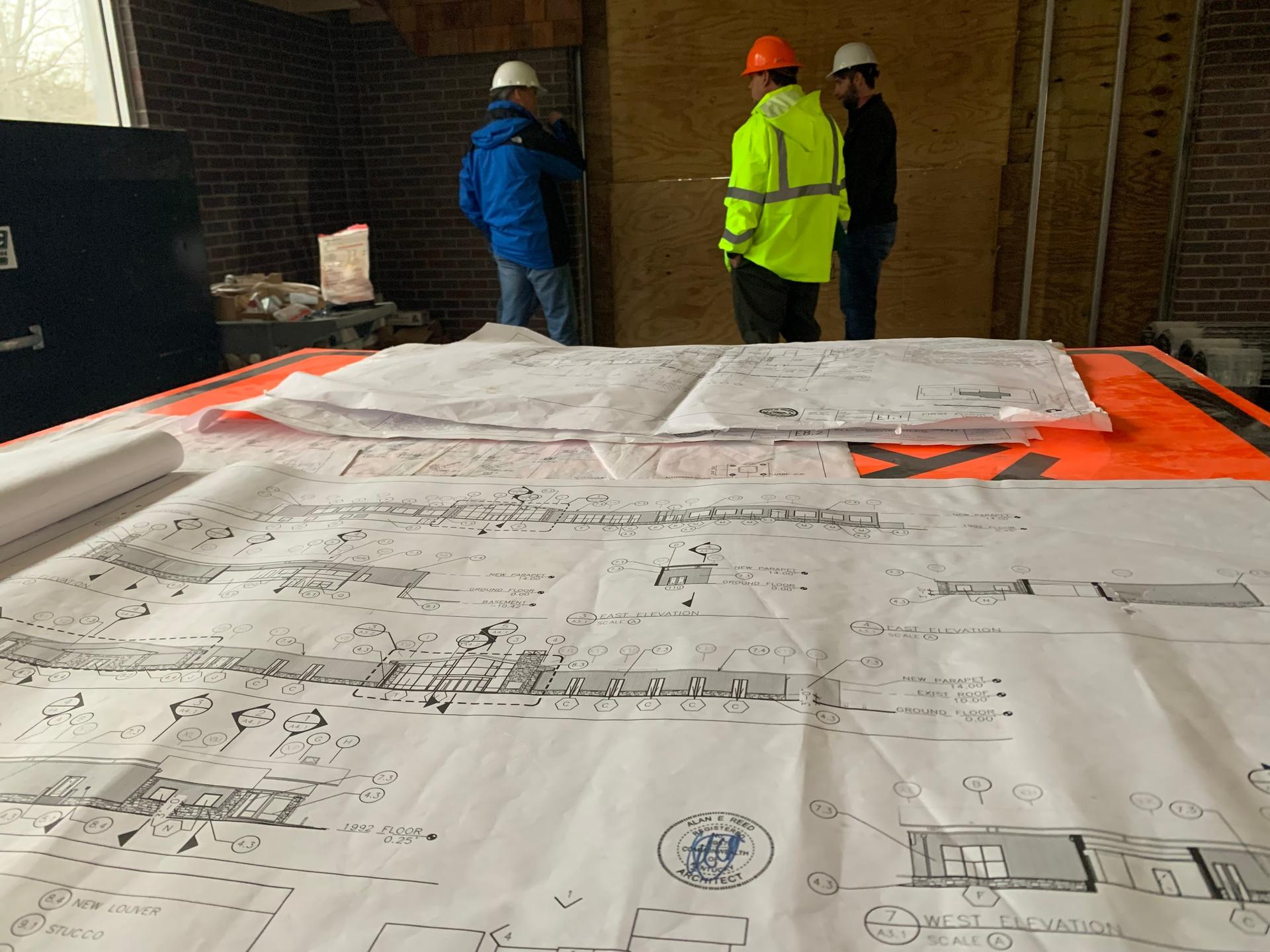 Plans de construction grand format étalés sur une table de chantier, ouvriers casqués au second plan
